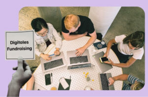 Hand hält Schild welches sagt digitales Fundraising. Im Hintergrund sitzen Menschen am Tisch vor Laptop.