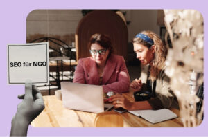 Hand hält Schild hoch welches sagt SEO für NGO. Hinten sitzen zwei Frauen und schauen in Laptop